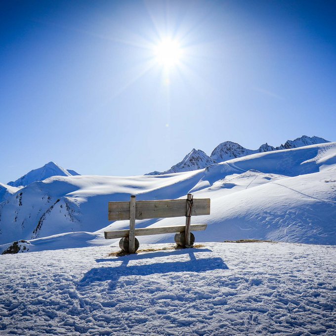 Urlaub im Kuschelhotel in Österreich Holzbank auf schneebedecktem Berg mit strahlender Sonne und klarem blauem Himmel