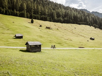 Looking for a hotel in Prutz? Two cyclists riding on a path through a green alpine meadow with small huts and forest
