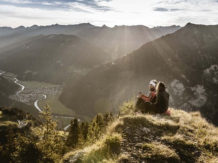 Looking for a hotel in Prutz? Two hikers sitting on mountain summit overlooking valley and river under sunlight rays