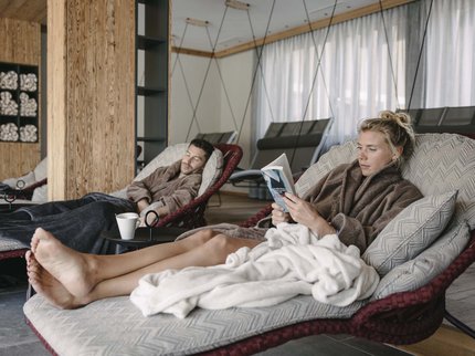 Ihr Urlaub im Sporthotel in Tirol Zwei entspannte Menschen in Bademänteln lesen und ruhen auf Sesseln in einem Spa.