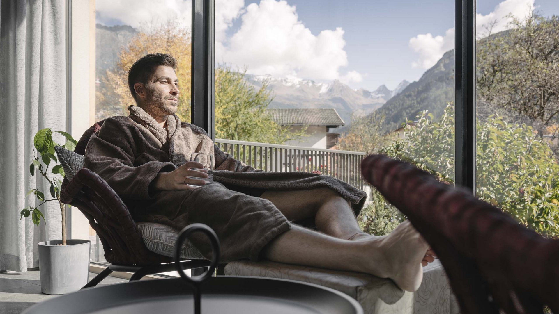 Looking for a hotel with spa in Tyrol? Man in bathrobe relaxing with drink by large window showing mountain view