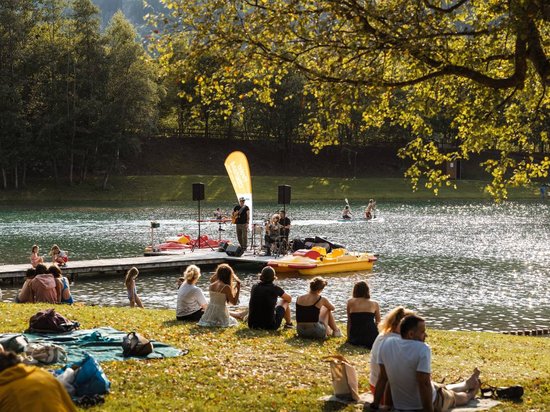 Echtes Kleinod aus dem Tiroler Oberland Menschen sitzen am Seeufer und hören Live-Musik auf einem Steg im Sonnenschein