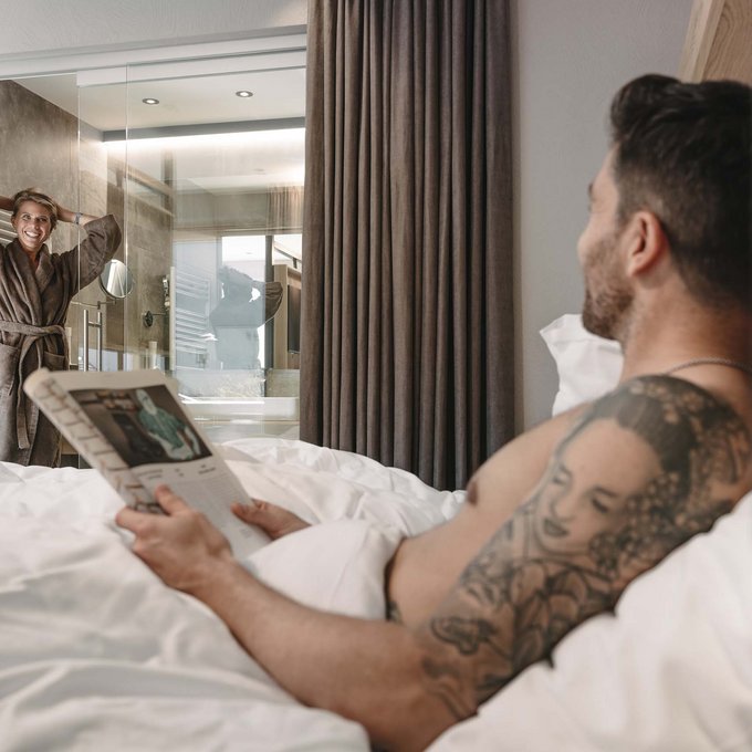 Urlaub im Kuschelhotel in Österreich Mann im Bett liest Zeitung, Frau im Bademantel im Badezimmer lächelt