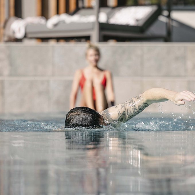 Outdoorpool Person mit Tattoo schwimmt im Pool, andere Person sitzt im Hintergrund