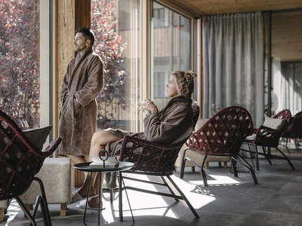 Urlaub im Kuschelhotel in Österreich Mann steht und Frau sitzt in Bademänteln am großen Fenster in einer modernen Lounge