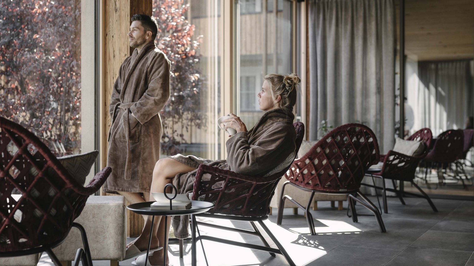 Looking for a hotel with spa in Tyrol? Man standing and woman sitting in bathrobes by large window in a modern lounge