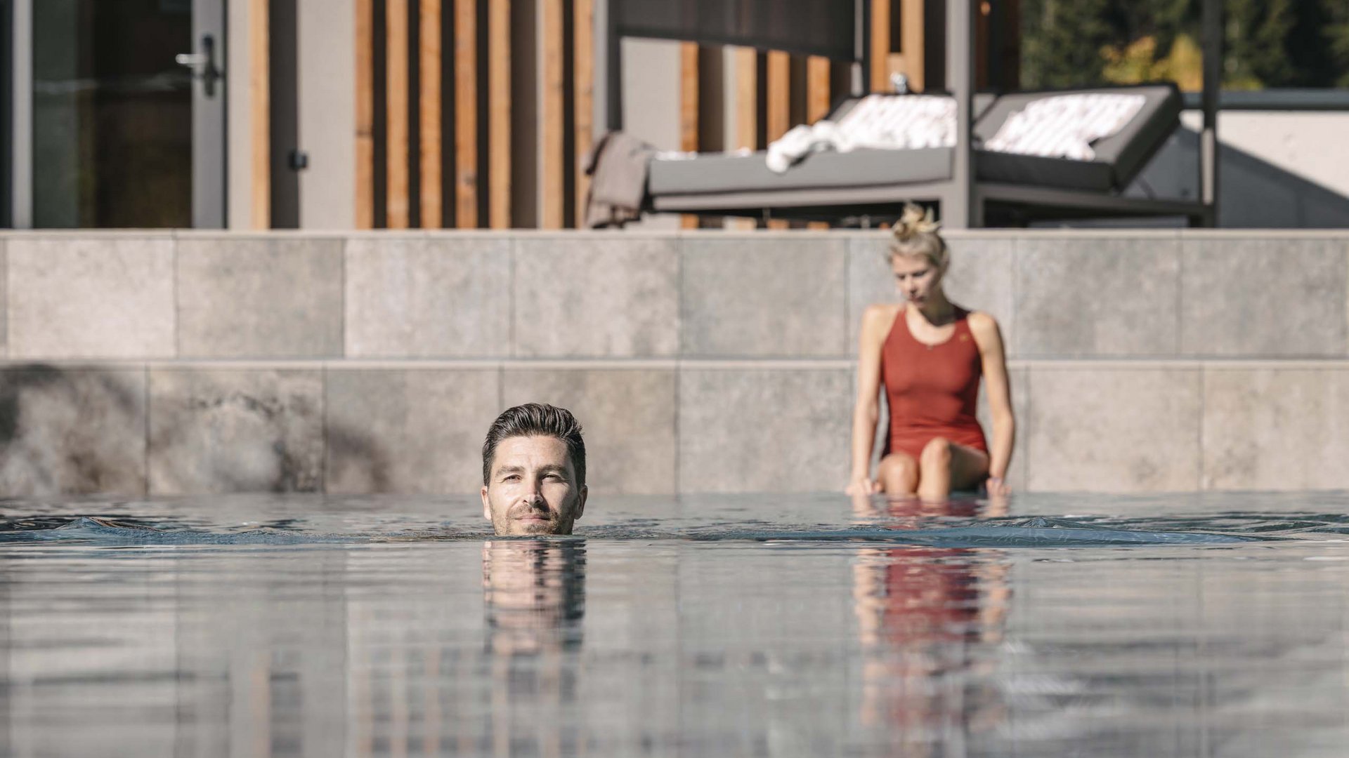 Looking for a hotel with spa in Tyrol? Man swimming in pool, woman sitting on edge wearing red swimsuit