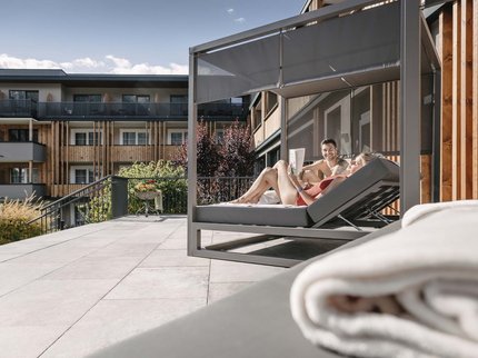Ihr Urlaub im Sporthotel in Tirol Paar entspannt auf Liege mit Sonnenschutz auf Terrasse eines modernen Gebäudes