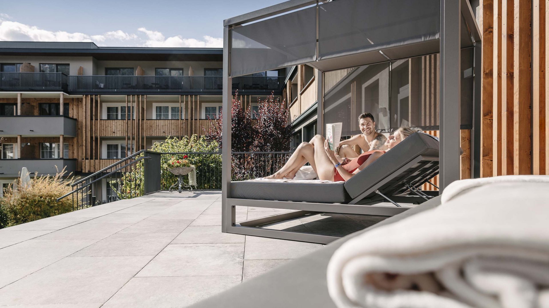 Looking for a hotel with spa in Tyrol? Couple relaxing on sunshade lounge chair on terrace of modern building