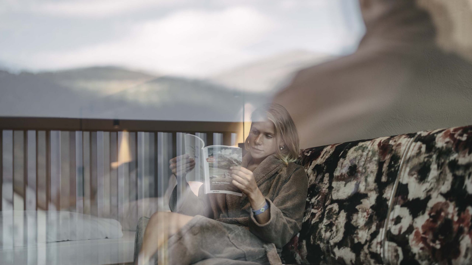 Your Tyrolean Oberland hotel Woman reading on floral sofa, photo taken through window with reflections