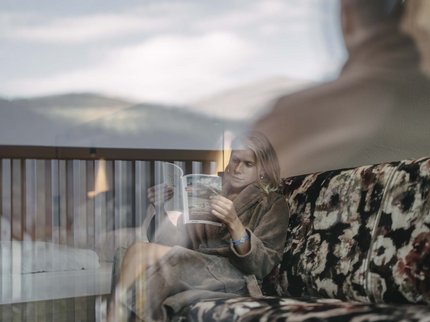 Urlaub im Kuschelhotel in Österreich Frau liest auf einem Sofa mit Blumendruck, durch ein Fenster mit Reflexionen fotografiert
